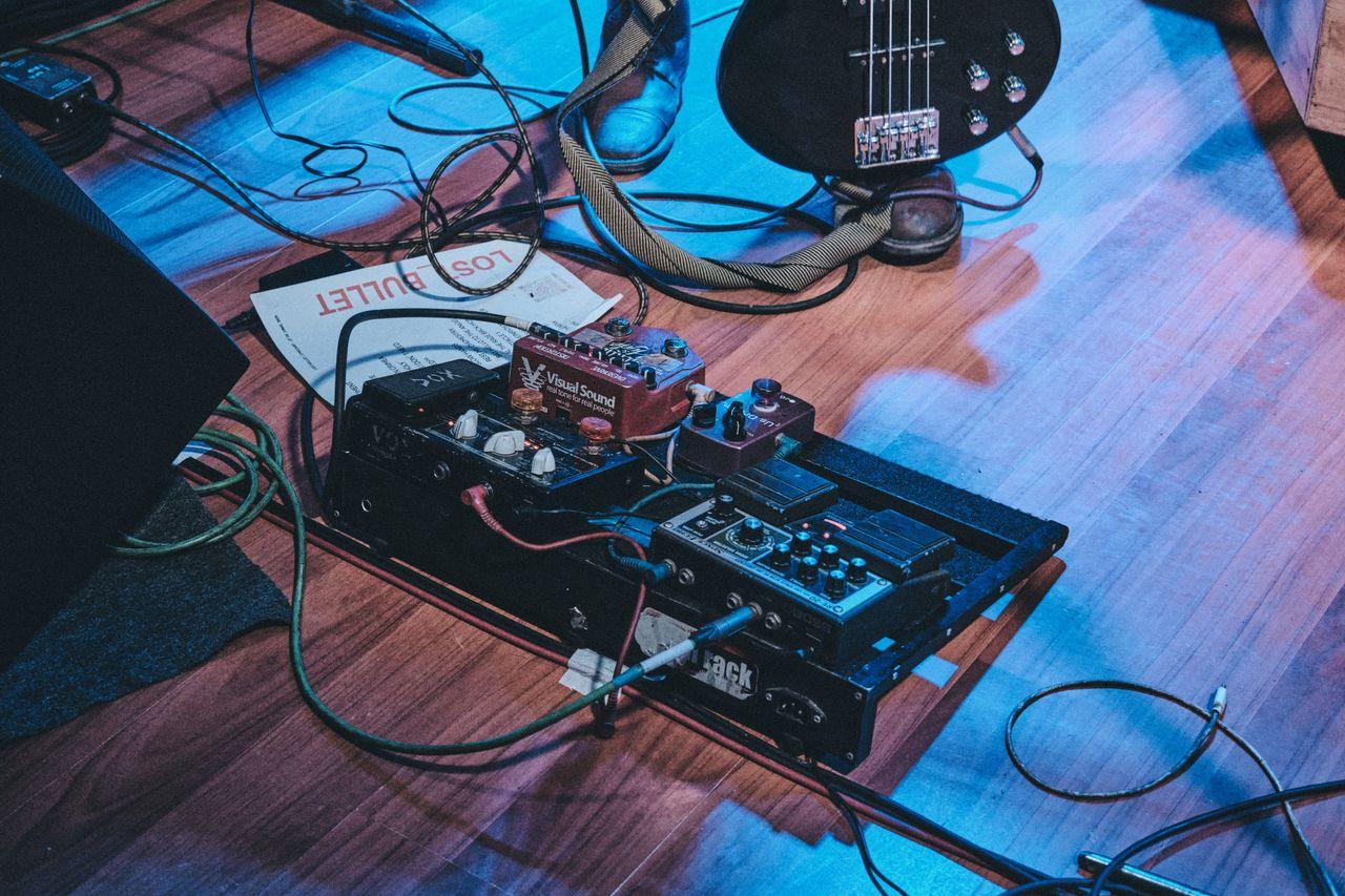 Offerings Close-up of a guitar pedal board on stage floor with a bass guitar and cables.